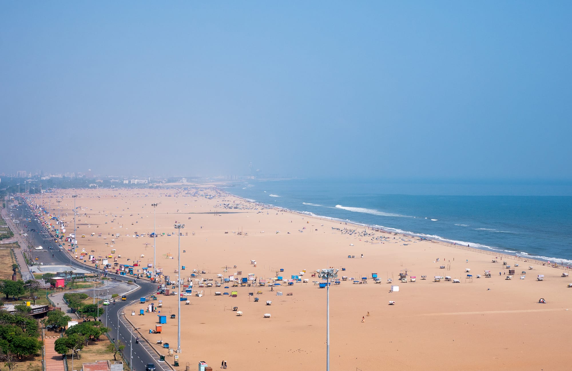 Aerial view of a wide sandy beach with sparse crowds and ocean waves, city buildings visible in the distance.